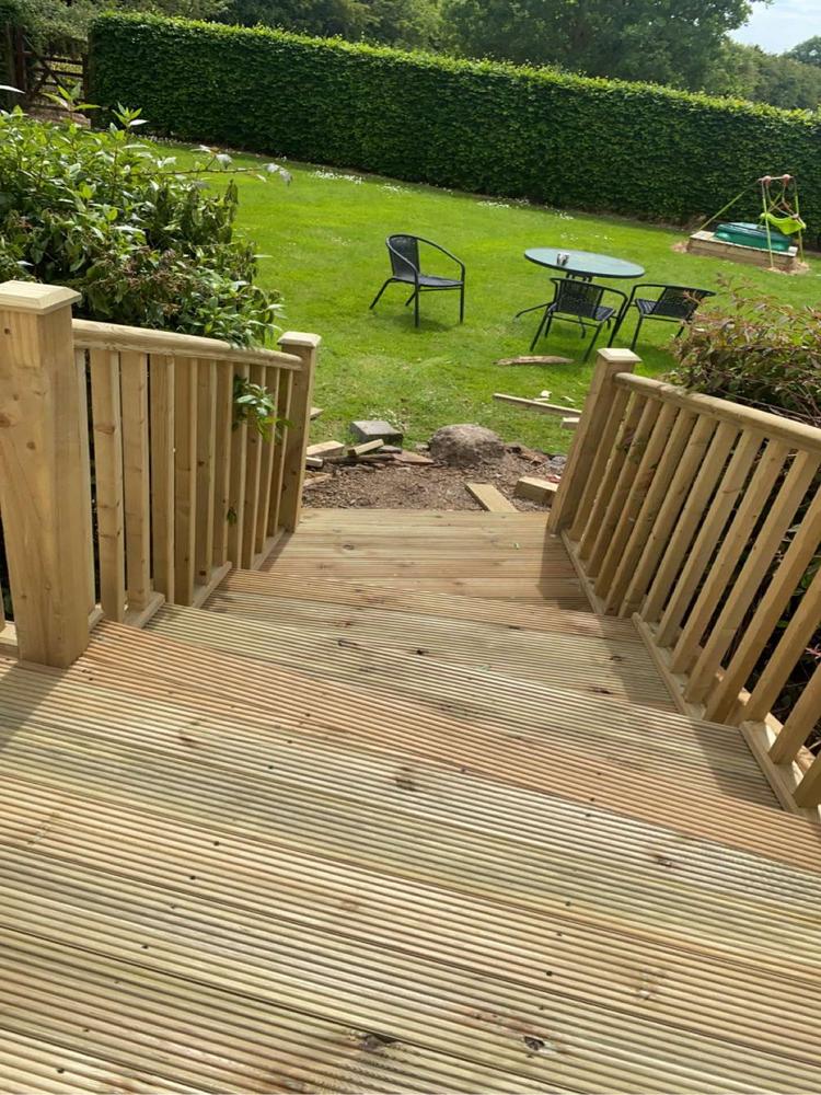 Wooden deck steps leading down to a garden with a lush hedge, lawn, outdoor table with chairs, and a children's swing set, showcasing timber craftsmanship and outdoor living.